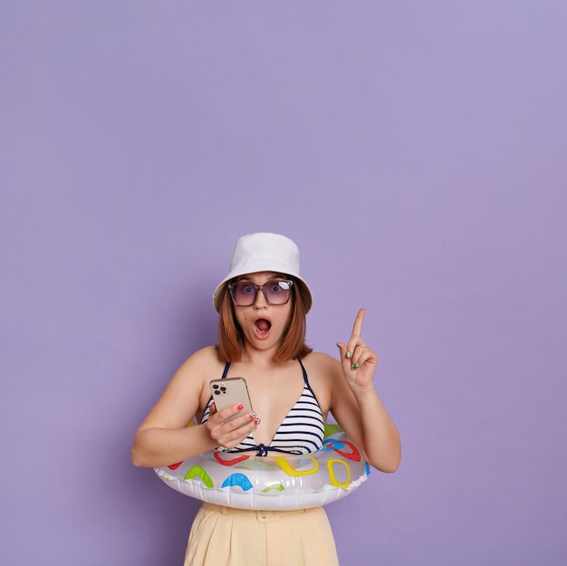 Imagem de mulher espantada chocada com anel de borracha vestindo panamá e maiô posando isolado sobre fundo roxo segurando o celular e apontando o dedo para o espaço da cópia tendo a ideia