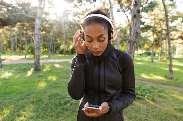 Imagem de mulher esportiva de 20 anos, vestindo um agasalho esportivo preto e fones de ouvido, usando um telefone celular enquanto caminha pelo parque verde