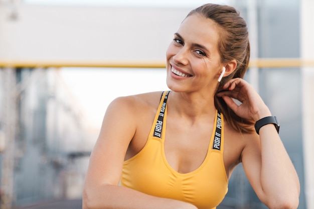 Imagem de mulher feliz em roupas esportivas com fones de ouvido sorrindo enquanto fica em pé sobre a grade ao ar livre