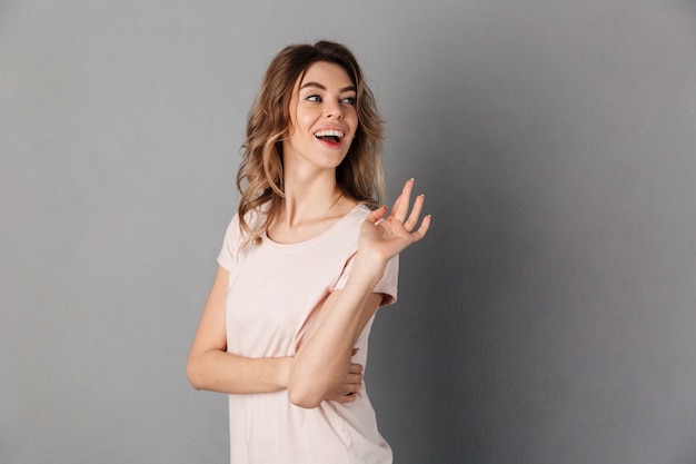 Imagem de mulher feliz surpresa em t-shirt acenando e olhando para trás sobre cinza