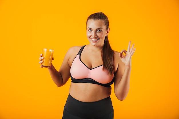 Imagem de mulher gordinha saudável em agasalho de treino sorrindo e segurando um copo com suco de laranja, isolado sobre fundo amarelo