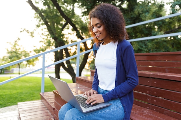 Imagem de mulher jovem africana feliz sentado ao ar livre no parque em degraus, usando o computador portátil.