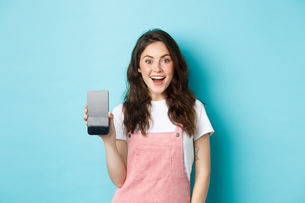 Imagem de mulher jovem e bonita parece surpresa e animada, mostrando a tela vazia do smartphone, seu logotipo ou aplicativo na tela do celular, em pé sobre um fundo azul.
