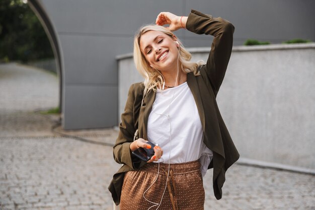 Imagem de mulher loira de 20 anos usando fones de ouvido sorrindo e usando smartphone enquanto caminha ao ar livre