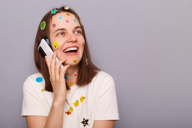 Imagem de mulher positiva sorridente coberta com adesivos engraçados posando isolada sobre fundo cinza falando no telefone inteligente e rindo espaço de cópia para propaganda