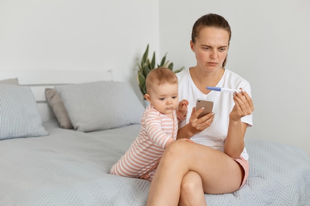 Imagem de mulher triste com tristeza expressando vestindo camiseta branca sentada na cama com a filha segurando telefone inteligente e teste de gravidez nas mãos sendo chateado com o resultado