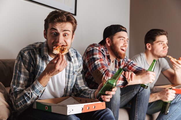 Imagem de três caras famintos comendo pizza com prazer, assistindo atentamente a partida de futebol em casa