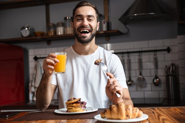 Imagem de um cara atraente dos anos 30 sentado à mesa e comendo enquanto toma o café da manhã em um apartamento moderno