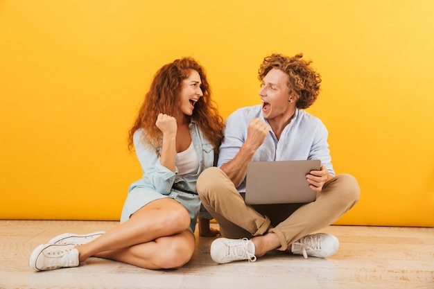 Imagem de um casal adorável e satisfeito, homem e mulher, sentados no chão e usando um laptop prateado, isolado sobre um fundo amarelo