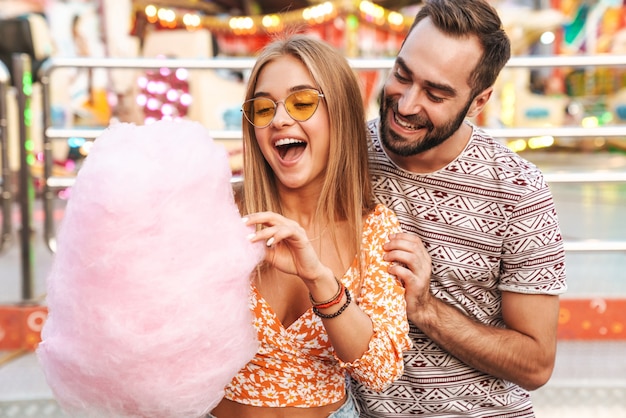 Imagem de um casal amoroso positivo sorridente, caminhando ao ar livre no parque de diversões, comer algodão doce.