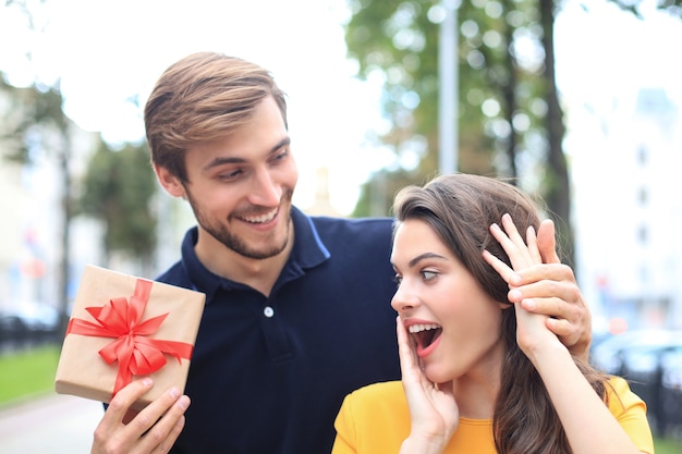 Imagem de um casal encantador e animado com roupas de verão, sorrindo e segurando a caixa de presente ao ar livre.