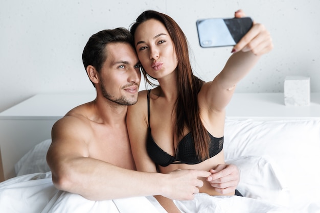 Imagem de um casal europeu, um homem e uma mulher, tirando uma foto de selfie juntos, enquanto está deitado na cama em casa ou apartamento de hotel