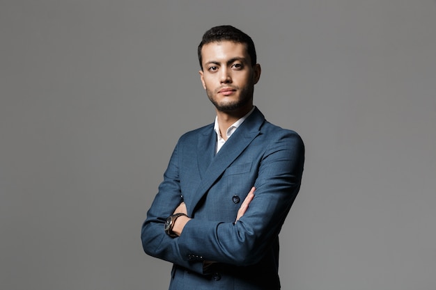 Imagem de um empresário árabe de 30 anos, barba por fazer, em terno formal, de pé com os braços cruzados, isolado sobre uma parede cinza