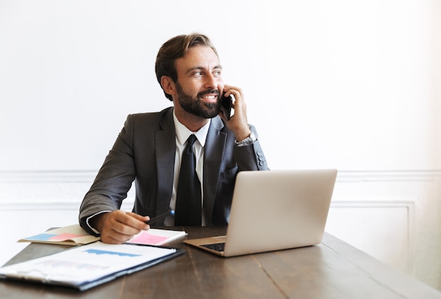 Imagem de um empresário satisfeito sorridente, vestindo um terno formal, falando ao celular enquanto trabalhava em um laptop no escritório