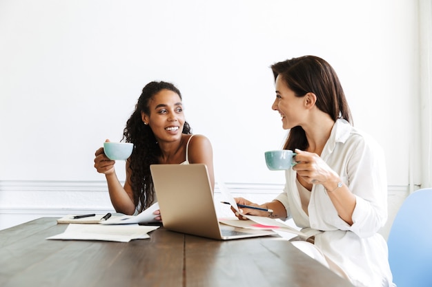 Imagem de um feliz incrível colegas de mulheres muito jovens no local de trabalho dentro de casa no trabalho de escritório com documentos em papel e computador portátil.