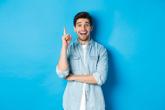 Imagem de um homem adulto bonito, tendo uma ideia, levantando o dedo e sorrindo animado, encontrou a solução, em pé contra um fundo azul