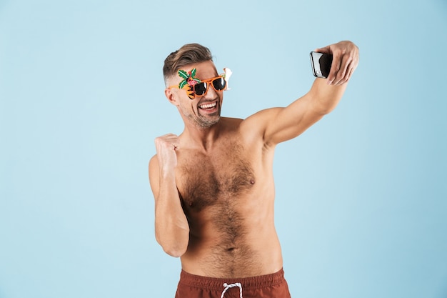 Imagem de um homem adulto feliz animado bonito em trajes de banho posando sobre uma parede azul, usando telefone celular, tire uma selfie.