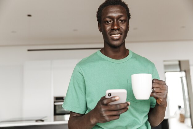 Imagem de um homem afro-americano otimista em roupas casuais segurando uma xícara de café e um smartphone em casa
