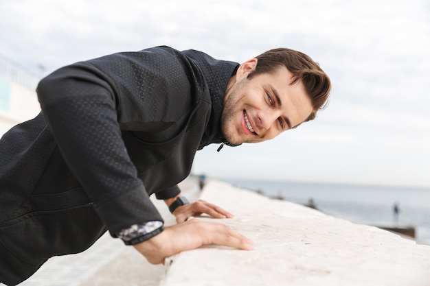 Imagem de um homem alegre de 30 anos em uma roupa esportiva preta, sorrindo enquanto caminhava pelo calçadão