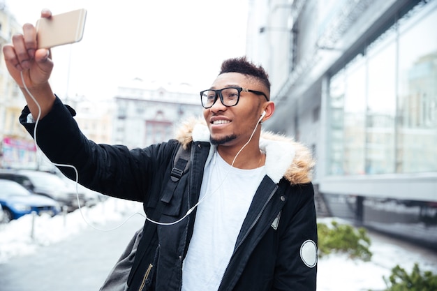 Imagem de um homem alegre segurando seu celular nas mãos e fazer uma selfie enquanto ouve música ao ar livre.