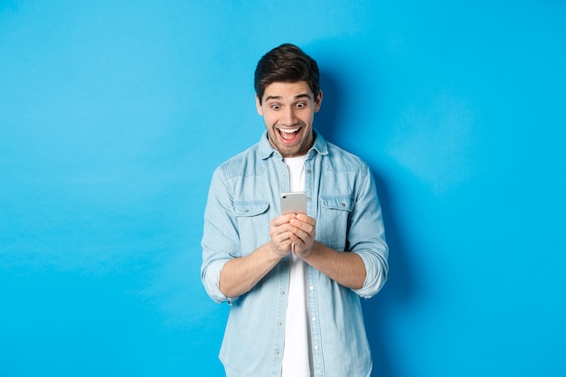 Imagem de um homem animado sorrindo enquanto olha para o celular, fazendo compras online no smartphone, em pé contra um fundo azul
