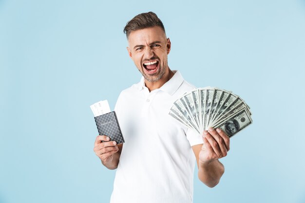 Imagem de um homem bonito, animado emocional gritando adulto posando sobre uma parede azul, segurando o passaporte com ingressos e dinheiro.