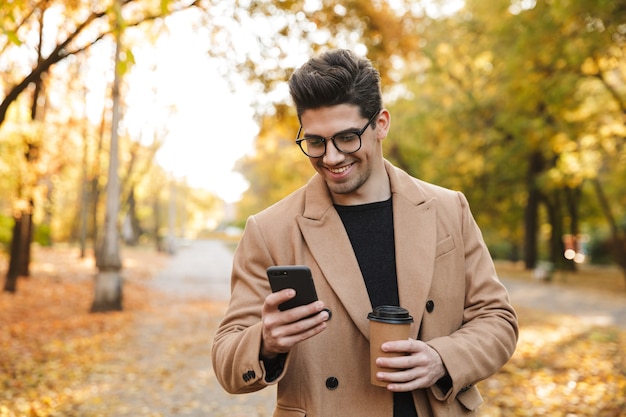 Imagem de um homem bonito e casual vestindo um casaco, usando o celular e sorrindo enquanto caminha no parque outono