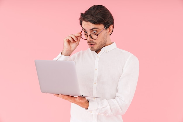 Imagem de um homem chocado bonito usando óculos, segurando e olhando para um laptop isolado