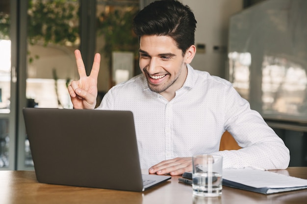 Imagem de um homem de 30 anos, alegre, do escritório, vestindo uma camisa branca, rindo e mostrando o símbolo da paz no laptop, durante videoconferência ou chamada