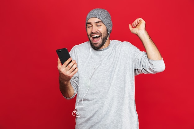 Imagem de um homem de 30 anos com a barba por fazer, em roupa casual, segurando um celular e ouvindo música com fones de ouvido isolados