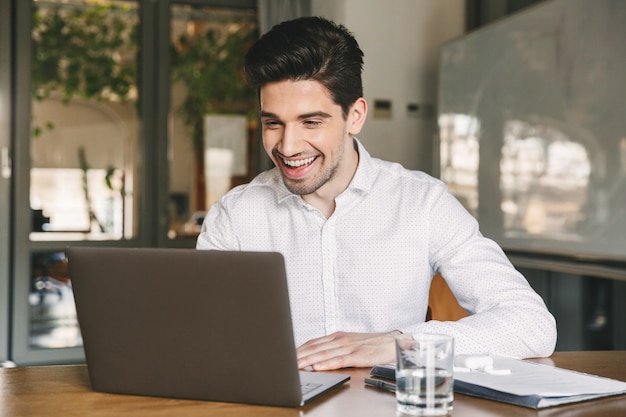 Imagem de um homem de negócios bonito de 30 anos vestindo uma camisa branca, rindo enquanto está sentado à mesa no escritório e olhando para o laptop