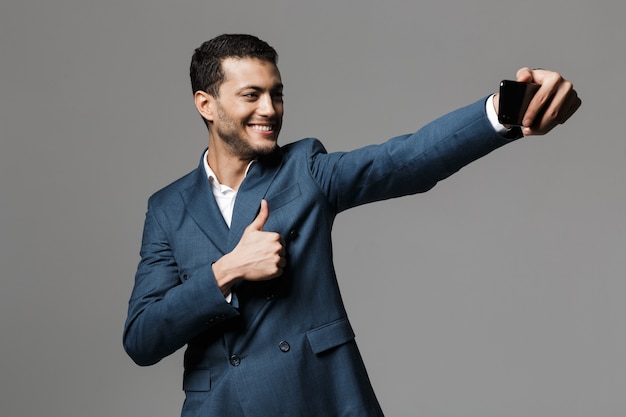 Imagem de um homem de negócios bonito tirar uma selfie pelo telefone móvel posando isolado sobre a parede de parede cinza.