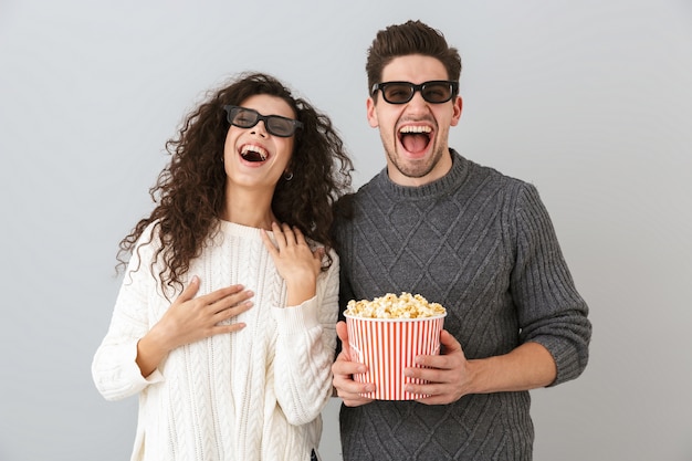 Foto imagem de um homem e uma mulher felizes usando óculos 3d segurando um balde com pipocas, isolado sobre uma parede cinza