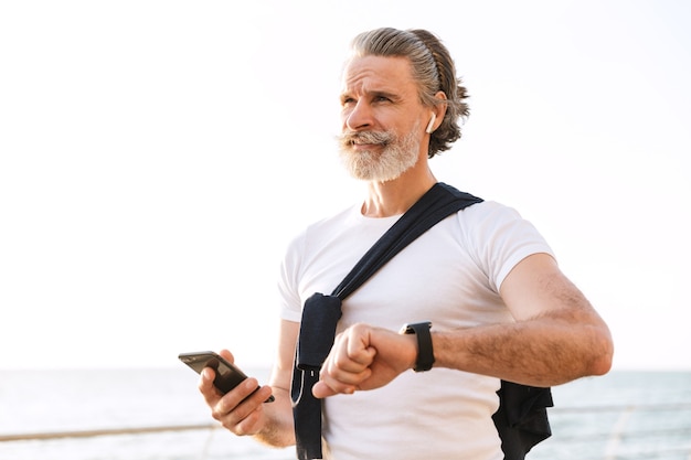 Imagem de um homem idoso satisfeito em roupas esportivas usando smartwatch e celular enquanto se exercita no calçadão pela manhã