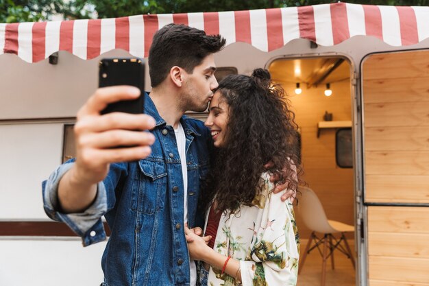 Imagem de um homem romântico beijando uma linda mulher e tirando uma selfie no celular perto de uma casa sobre rodas ao ar livre