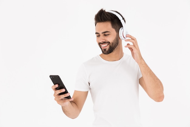 Imagem de um homem sorridente com a barba por fazer ouvindo música no celular e fones de ouvido isolados na parede branca