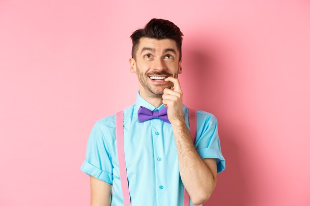 Imagem de um homem sorridente fazendo escolha, parecendo sonhador e feliz, roendo a unha com expressão tentada, querendo algo, em pé sobre um fundo rosa.