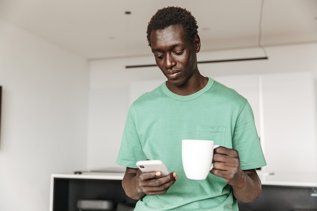 Imagem de um jovem afro-americano bonito em roupas casuais segurando um smartphone e uma xícara de café em casa