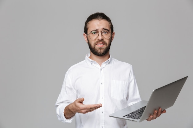 Imagem de um jovem barbudo bonito confuso posando isolado sobre uma parede cinza usando óculos, usando o computador portátil.