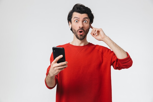 Imagem de um jovem bonito animado posando sobre uma parede branca, ouvindo música com fones de ouvido, usando telefone celular.