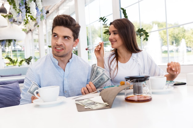 Imagem de um jovem casal amoroso confuso descontente, sentado no café, segurando cheque e dinheiro.