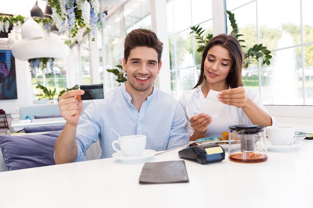 Imagem de um jovem casal amoroso feliz sentado no café, segurando o cartão de crédito e cheque.