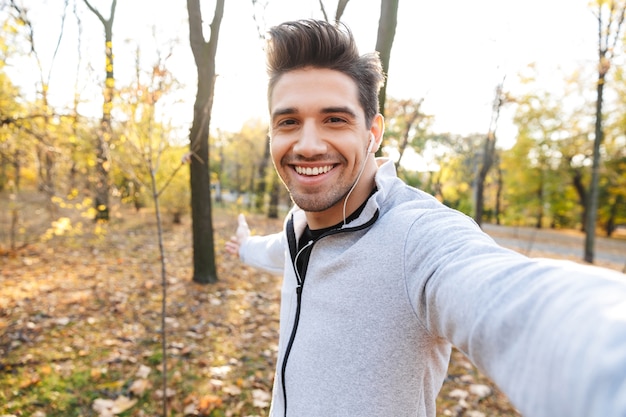 Imagem de um jovem desportista bonito ao ar livre no parque ouvindo música com fones de ouvido tirar uma selfie pela câmera.