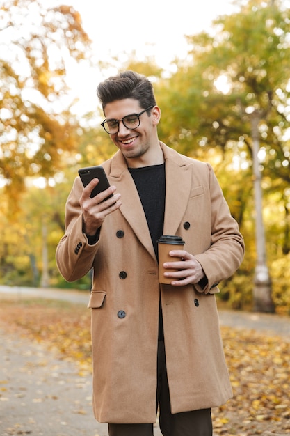 Imagem de um jovem feliz vestindo um casaco, digitando em um smartphone e sorrindo enquanto caminha no parque outono