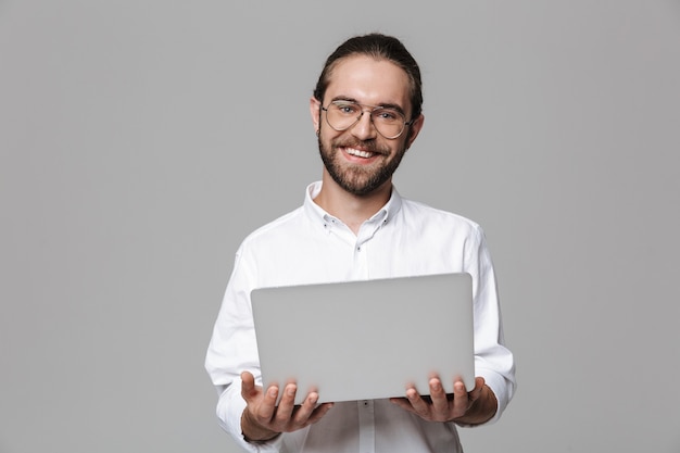 Imagem de um jovem homem barbudo bonito positivo posando isolado sobre uma parede cinza usando óculos, usando o computador portátil.