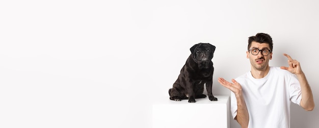 Imagem de um jovem intrigado sentado perto de um lindo pug preto mostrando tamanho pequeno e encolhendo os ombros