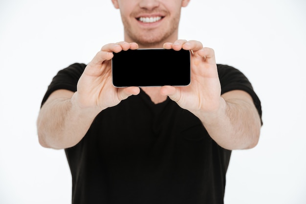 Imagem de um jovem sorridente vestido com uma camiseta preta em pé sobre um fundo branco, mostrando o visor de seu telefone para a câmera.