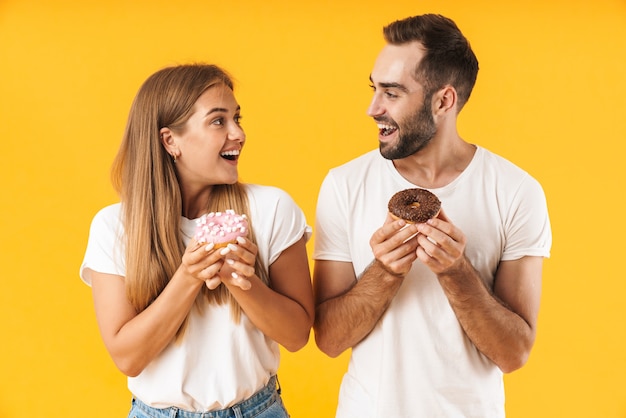 Foto imagem de um lindo casal, homem e mulher, sorrindo, segurando rosquinhas doces juntas isoladas sobre uma parede amarela