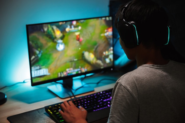 Imagem de um menino adolescente imerso jogando videogame no computador em um quarto escuro, usando fones de ouvido e um teclado colorido com luz de fundo