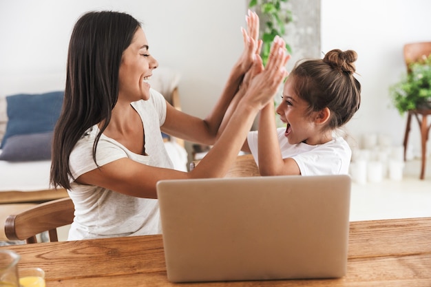 Imagem de uma alegre mulher de família e sua filha dando mais cinco e usando um laptop juntos, enquanto estão sentados à mesa no apartamento